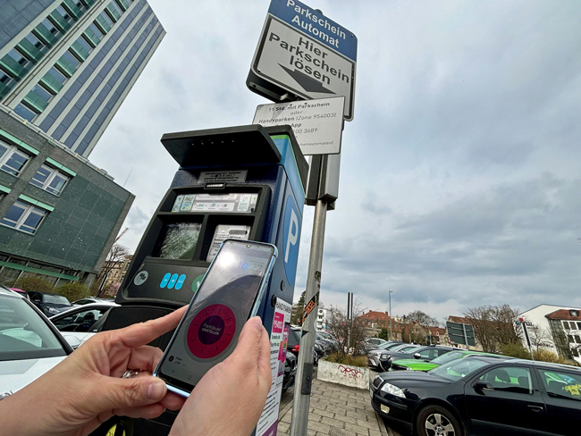 Vor einem Parkscheinautomaten wird ein Handy mit einer geöffneten App auf dem Bildschirm hochgehalten. Im Hintergrund sieht man parkende Autos.