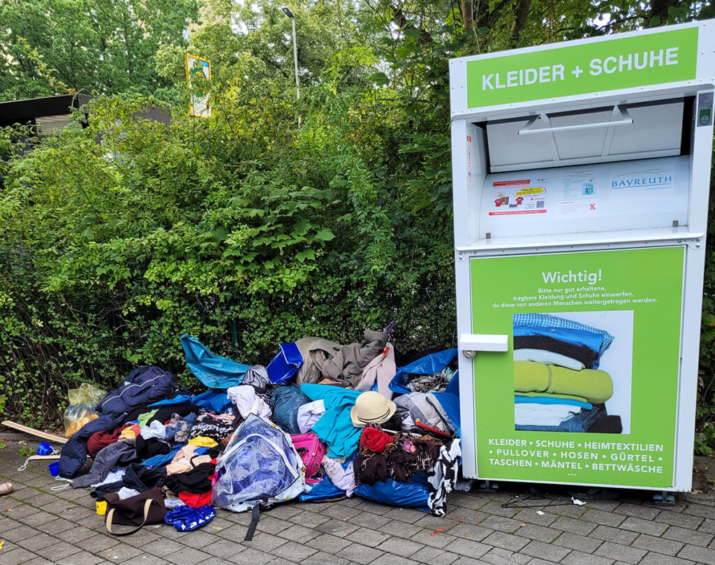 Ein Haufen Klamotten liegt zerstreut am Boden neben dem Altkleidercontainer